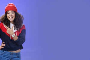 stylish and sociable friendly looking attractive woman with curly hair in warm beanie pointing at upper left corner laughing from joy and amazement having fun posing happily over blue background